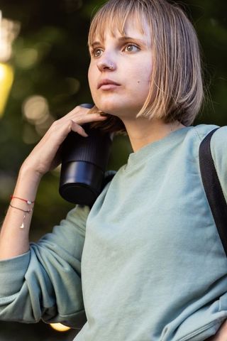 Young woman holding travel mug carrying backpack looking contemplative in park golden hour
