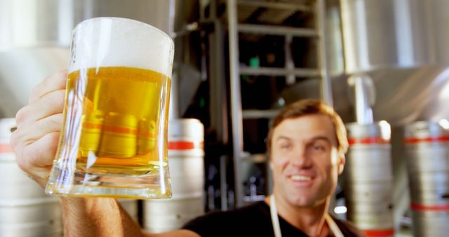 Professional brewer proudly holding a glass of freshly brewed beer in a brewery with industrial equipment in the background. Perfect for scenes depicting the brewing process, appreciation of craft beer, and showcasing joyful professionals in the alcohol industry.
