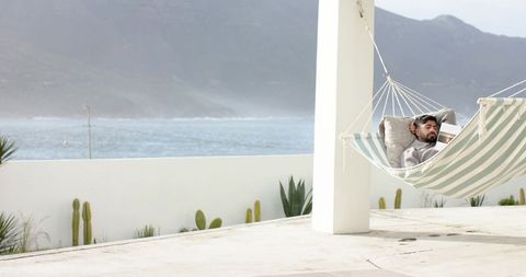 Indian man relaxing in striped hammock on coastal terrace reading book with ocean view