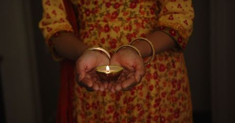 An Indian woman wears vibrant traditional attire and gracefully holds a lit diya. The flickering light signifies joy, spiritual harmony, and the celebration of Diwali, known as the festival of lights. The scene epitomizes cultural richness and the timeless tradition of lighting diyas as an element of positivity and hope. Ideal for use in articles about Indian festivals, spirituality, cultural practices, and traditional attire associated with festivals. Inspire with images of hope and cultural rituals seen in Diwali celebrations across communities.