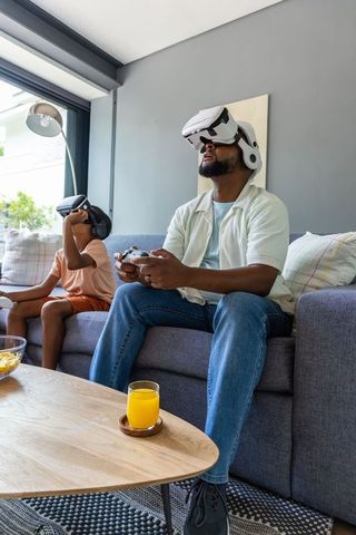 African american dad and son wearing vr headsets playing games on living room couch