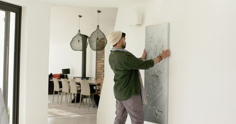 Young man hanging large abstract canvas on white wall in modern open-plan living space