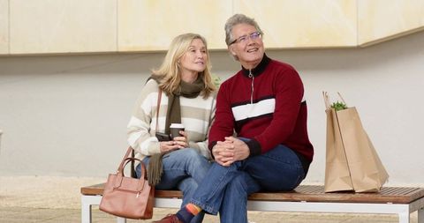 Senior couple relaxing on bench, holding coffee and smartphone, shopping bags by side