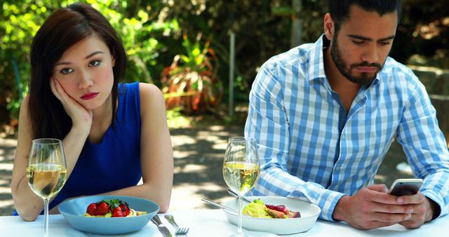 Woman sitting bored and frustrated while man distracted by his smartphone at an outdoor restaurant. The scene highlights modern-day relationship issues, lack of communication, and the impact of technology on personal interactions. Suitable for use in articles about relationships, social interaction, technology's influence on daily life, and dining experiences.