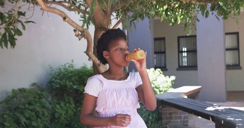 Teen girl drinking orange juice while standing under tree in sunny courtyard for summer refreshment