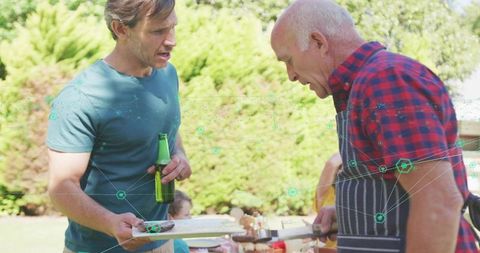 Senior man grilling skewers while younger man holding beer bottle at backyard family cookout