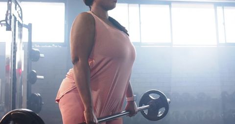 African american woman lifting barbell in gym wearing pink activewear and fitness tracker