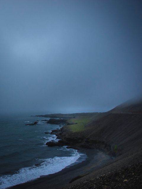 Moody coastal cliff with foggy atmosphere and waves crashing against rocks during dusk. Perfect for themes of nature, solitude, meditation, and dramatic landscapes. Could be used in travel promotion, environmental studies, or backgrounds for presentations.