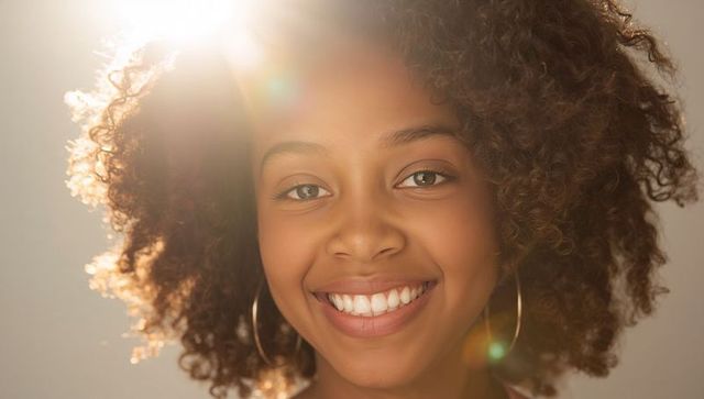 Smiling young woman with voluminous curly hair backlit by warm lens flare and hoop earrings