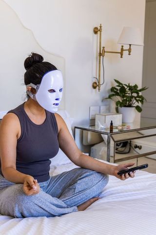 Woman using led facial mask on bed holding smartphone for at-home skincare and relaxation