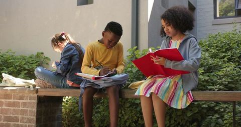 Teen students studying outdoors on campus bench writing notes in notebooks and red folder