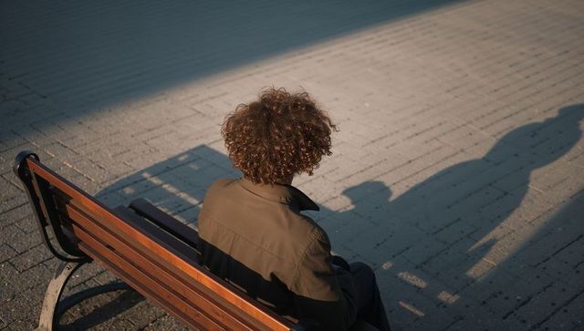 Curly-haired man sitting on bench at golden hour casting long shadow across urban plaza