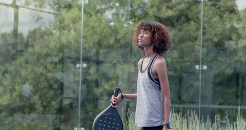 Young african american woman holding padel paddle standing on outdoor glass court
