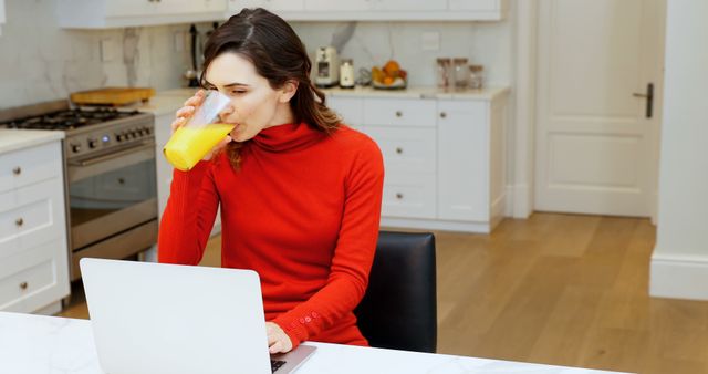 Woman sits at kitchen table, drinking orange juice and working on a laptop. She wears a red sweater, suggesting a casual home setting. Useful for illustrating remote work, multitasking, morning routines, or healthy living concepts.