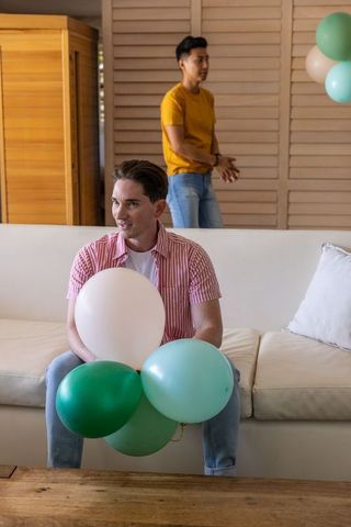 Male friends relaxing on sofa holding pastel balloons at casual home celebration