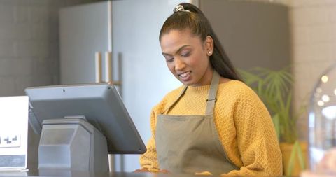 Indian barista tapping pos touchscreen at cafe counter wearing mustard sweater and apron
