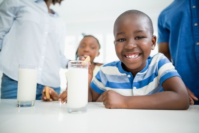 Boy smiling while enjoying cookies and milk in a home kitchen. Family members in the background. Ideal for use in advertisements for dairy products, family-oriented brands, or healthy snack promotions. Can also be used in articles about childhood nutrition, family bonding, or home life.