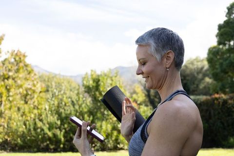 Senior woman preparing for outdoor yoga checking smartphone while carrying rolled mat on lawn
