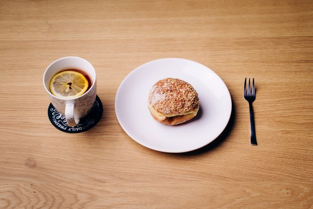 Cup of black tea with a lemon slice served on a wooden table next to a plate with a pastry bun and a fork. Perfect for articles, promotions, or visuals related to breakfast, afternoon tea, cozy setups, homemade food, and simple meals.