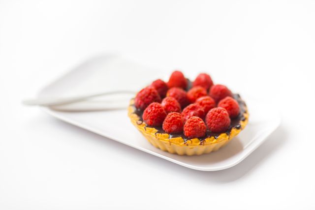 View of a delectable mini raspberry tart on a white plate, accompanied by a utensil. Perfect for using in websites or promotions related to desserts, pastries, bakery product advertising, gourmet websites, food blogs, or any content highlighting culinary delights.