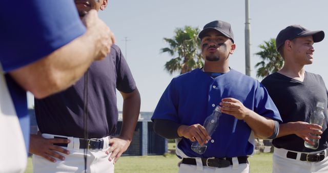 Group of baseball players pausing game to hydrate and discuss strategy. Players hold clear water bottles, appear engaged in conversation. Palm trees in background suggest warm, sunny day. Ideal for illustrating teamwork, sportsmanship, break time in sports activities, or hydration during physical exertion.