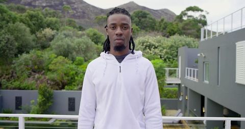 Young man standing on modern balcony wearing white hoodie overlooking garden and hillside