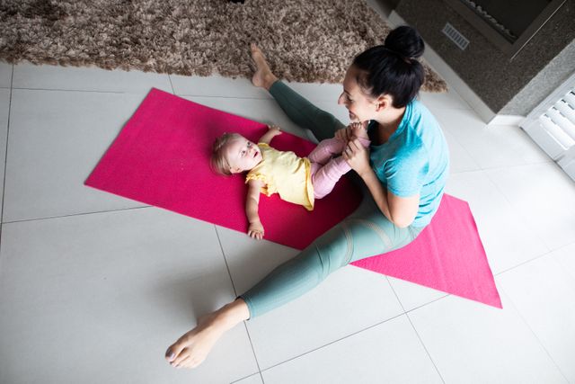 Mother engaging in a home workout while interacting with her baby daughter on a yoga mat. Ideal for use in articles or advertisements about home fitness, parenting, family bonding, and maintaining a healthy lifestyle during quarantine or lockdown.