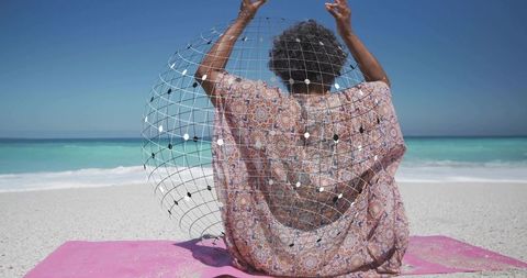 Senior woman sitting on turquoise beach raising arms with wireframe globe wearing patterned kaftan