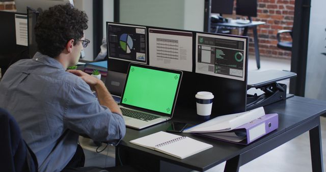 Business professional working at a desk with multiple computer screens displaying data and charts. An open laptop with a green screen for easy editing is also visible. A notebook with pen, coffee cup, smartphone, and organized files on the desk reflect a productive and organized workplace. Ideal for portraying modern office environments, data-driven workplace, and business productivity.