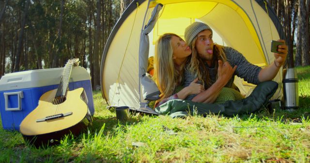 Young couple camping in a forest, taking a playful selfie inside a tent. The surrounding grassy area features sunlight streaming through trees, a blue cooler, and an acoustic guitar, creating a fun and relaxed outdoor atmosphere. Perfect for use in ads related to outdoor activities, camping gear promotions, vacation planning, and lifestyle blogs.