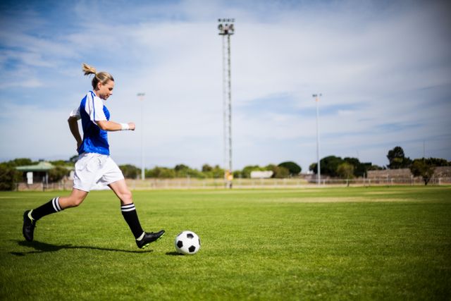 Female soccer player in action, practicing on a green soccer field. She is about to kick a ball, wearing a blue and white uniform. This can be used for sports training articles, women's soccer promotion, athletic gear advertisements, and content about outdoor activities.