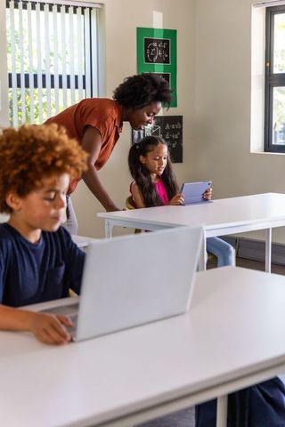 African American teacher guiding students using tablet and laptop in bright classroom
