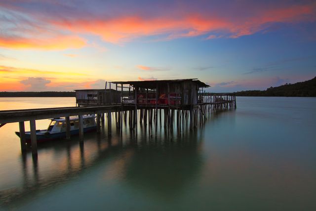 Calm tropical sunset with a wooden hut on stilts standing over calm water, and a boat docked at a wooden pier. The sky is painted with vibrant oranges, pinks, and blues, reflected in the water below. Suitable for promoting travel destinations, vacation resorts, or relaxation retreats. Ideal for background images in websites or social media campaigns aiming to depict peaceful and scenic seaside views.