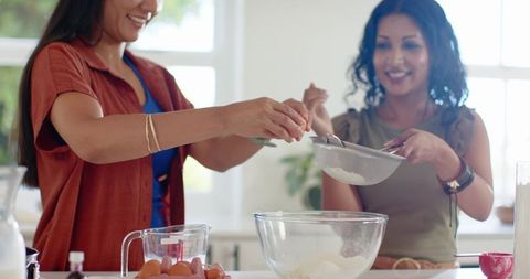Friends baking together cracking eggs and sifting flour in sunlit home kitchen