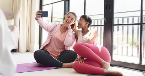 Two friends taking selfie after home workout on yoga mats by sunlit balcony window