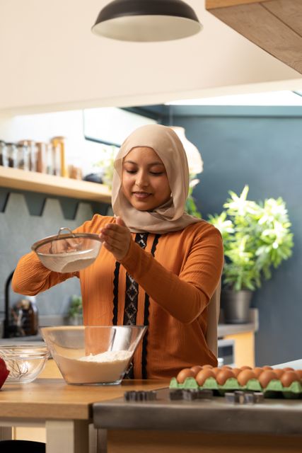 Smiling woman in hijab baking in a modern kitchen, mixing ingredients in a bowl. Ideal for use in articles or advertisements about home cooking, inclusivity, domestic life, and leisure activities. Perfect for promoting kitchen appliances, cooking classes, or lifestyle blogs.