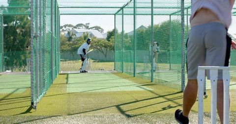 Coach walking behind batsman in cricket nets during outdoor turf training session