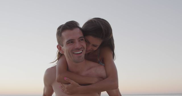 This stock photo depicts a joyful couple enjoying a romantic moment during a piggyback ride on a beach at sunset. The image emphasizes love, happiness, and togetherness and can be used in promotions for travel agencies, romantic getaways, vacation advertisements, and relationship articles.