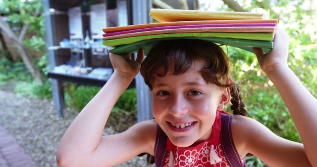 Young girl smiling while holding colorful paper folders on her head. She is outdoors with nature in the background. Perfect for use in educational materials, children's activities, summer camp promotions, creative workshops, or any content related to fun and learning.