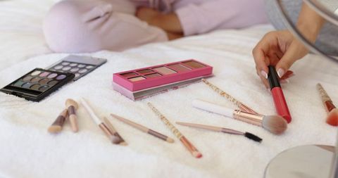 Holding coral lip gloss over pastel eyeshadow palettes and makeup brushes on plush white bedspread