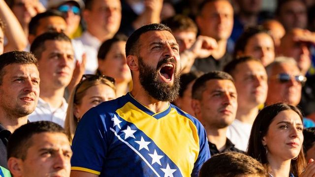 Bearded fan cheering in blue yellow jersey with white star sash in sunlit stadium crowd