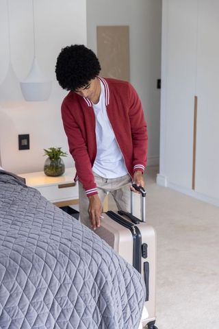 Young traveler placing beige rolling suitcase beside bed in modern minimalist bedroom