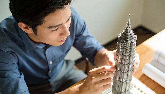 Asian architect assembling detailed skyscraper model on desk with blueprints and plans