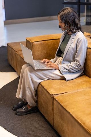 Young professional woman working on laptop on tan leather modular sofa in modern office lounge