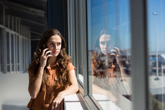 Businesswoman talking on mobile phone while looking through window at office. Ideal for use in business communication, corporate environments, technology in the workplace, and professional settings. Can be used in articles, blogs, and marketing materials related to business and professional life.