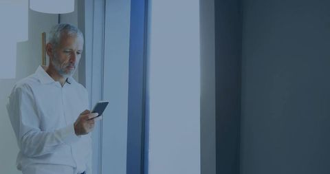 Senior businessman checking smartphone by office window with minimalist daylight workspace