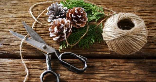 A rustic crafting scene is captured with pine cones, greenery, scissors, and twine on a wooden surface. It evokes a sense of creativity and the handmade charm of DIY projects or holiday decorations.