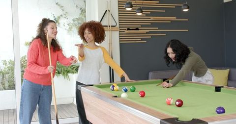 Diverse female friends playing billiards in modern home game room with green felt table