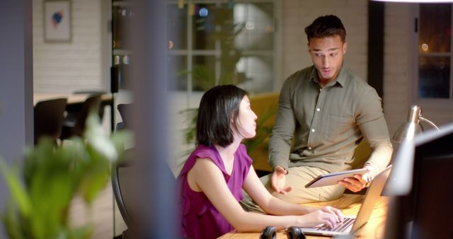 Professional man showing information on tablet to female co-worker working on laptop in modern office at night. Ideal for themes such as teamwork, collaboration, business discussions, and focused work in an office environment.