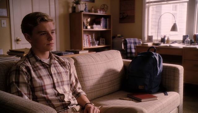 Teenage male sitting on dorm couch gazing thoughtful student with backpack, desk, books
